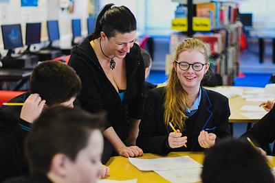 A writer standing beside a smiling pupil writing at a class desk